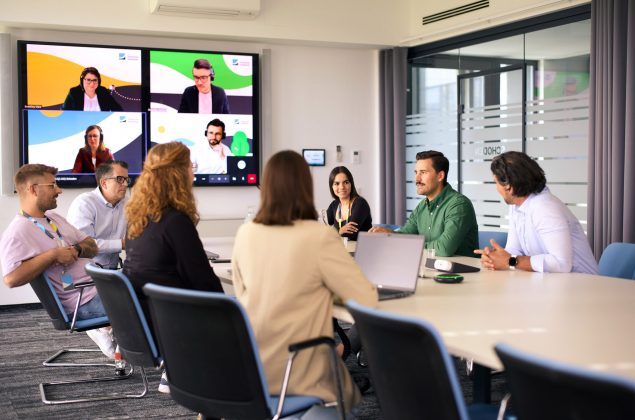 Sieben Personen an einem Konferenztisch, im Hintergrund ein großer Monitor mit zugeschalteten Kollegen